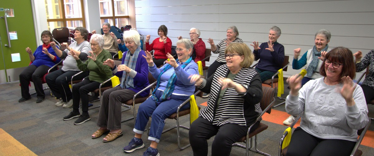 A group of older people sitting in chairs look happy as they do exercises.